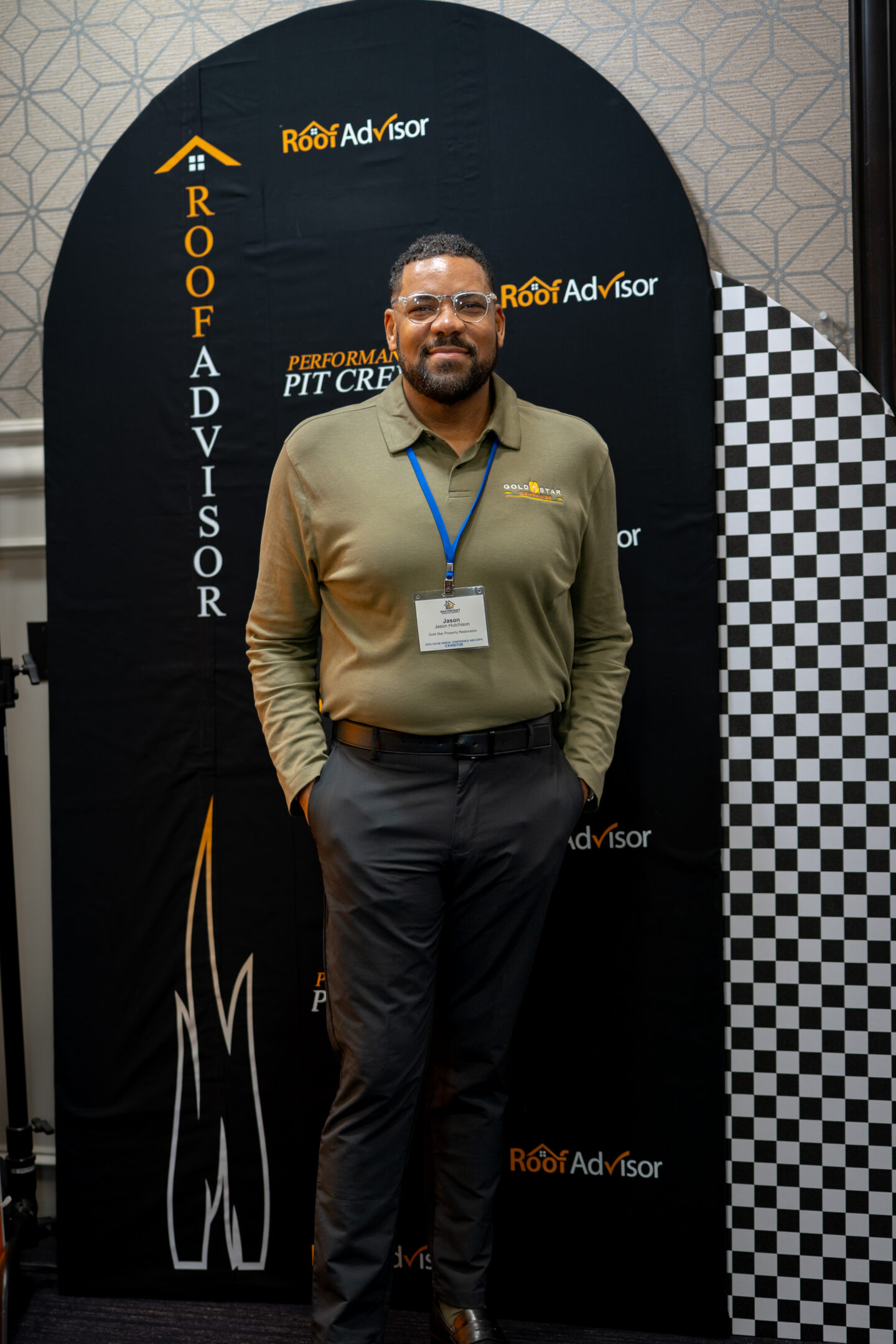 CAI202601: Man posing in front of a Roof Advisor backdrop. He is wearing glasses, a polo shirt, slacks, and a name tag. Professional headshot at event.