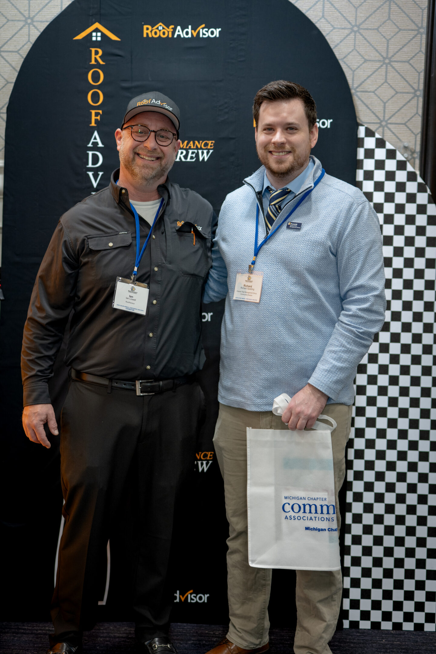 Two men pose at Roof Advisor event CAI2026010, showcasing performance crew. One wears a Roof Advisor hat. Michigan Chapter Comm Associations bag visible.
