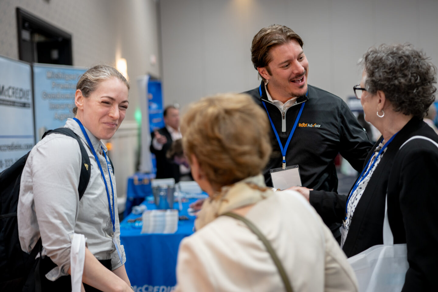 CAI2026011: Three people are engaged in conversation at a convention. One man wears a Roof Advisor jacket. The setting appears to be a business expo.