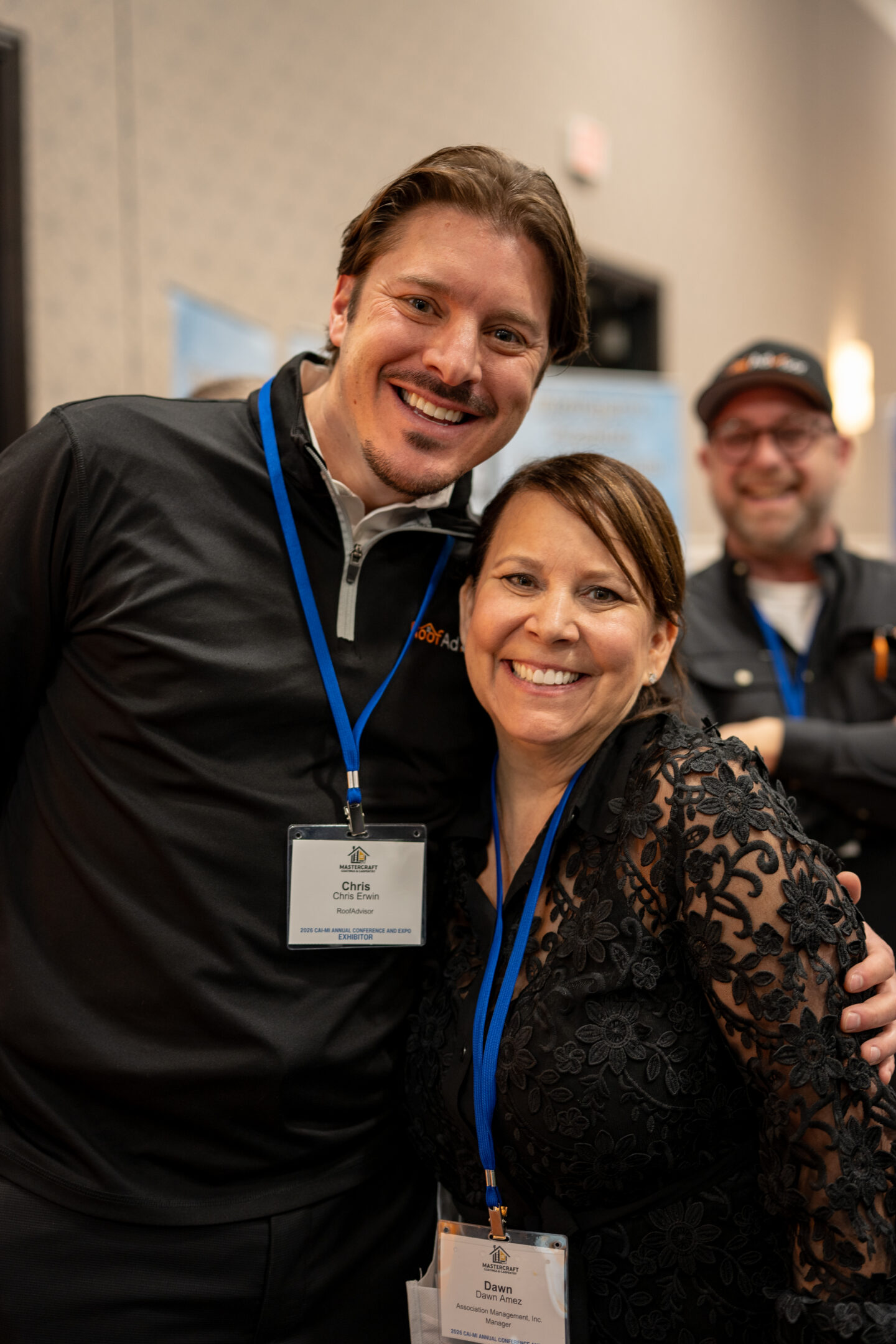CAI2026013.jpg: Two smiling professionals, Chris and Dawn, pose at a conference, wearing badges. Another attendee smiles in the background.