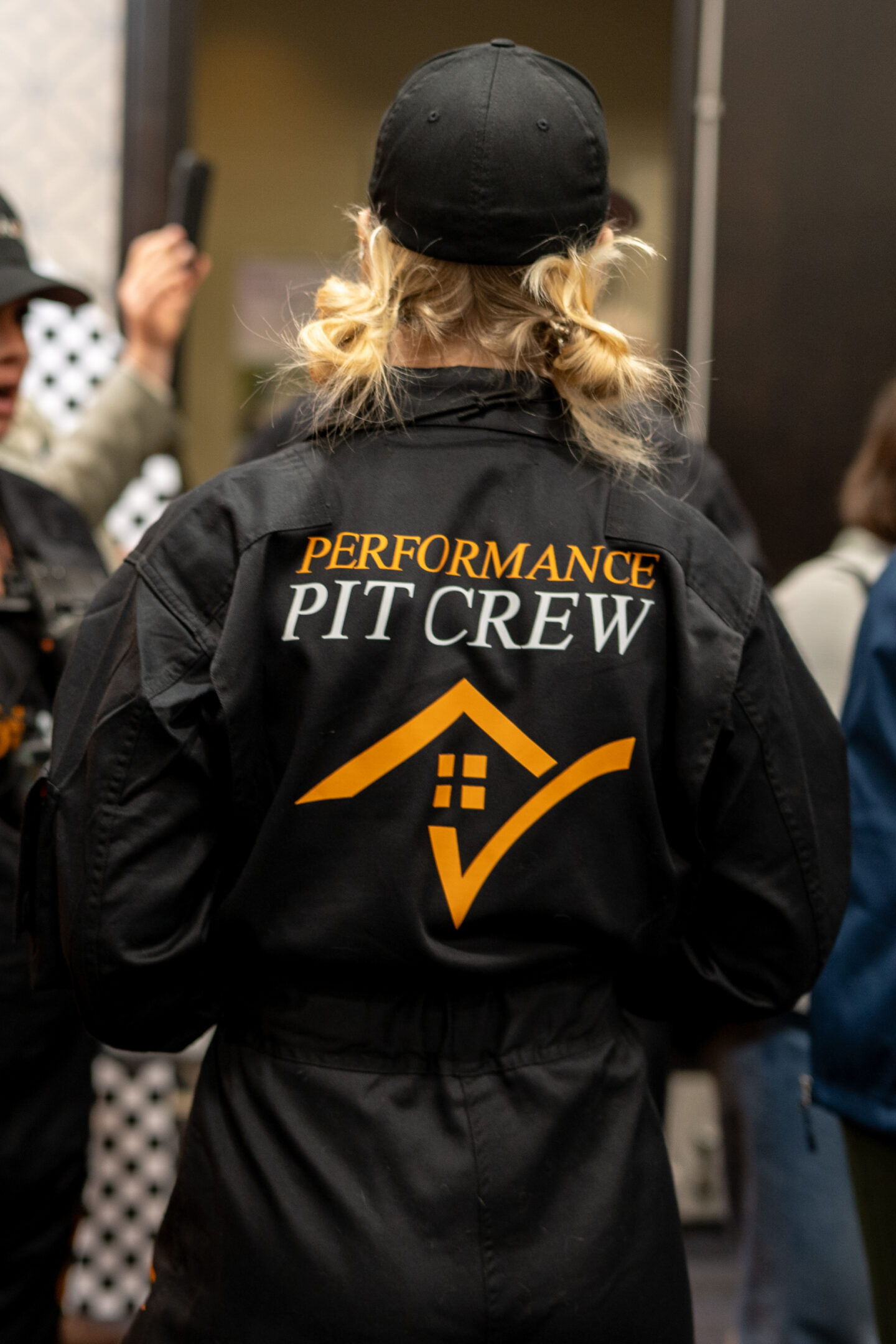 CAI2026014: Back view of a person wearing a black "Performance Pit Crew" jacket with an orange house logo and check mark, indicating performance excellence.