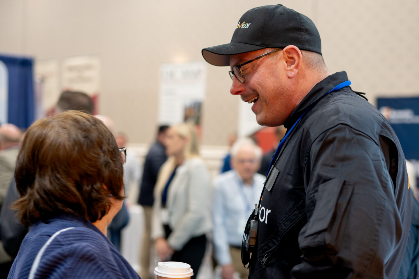 CAI2026016: Man in Advisor cap conversing at a conference. Networking event with attendees in background, engaging in discussions. Business meeting atmosphere.