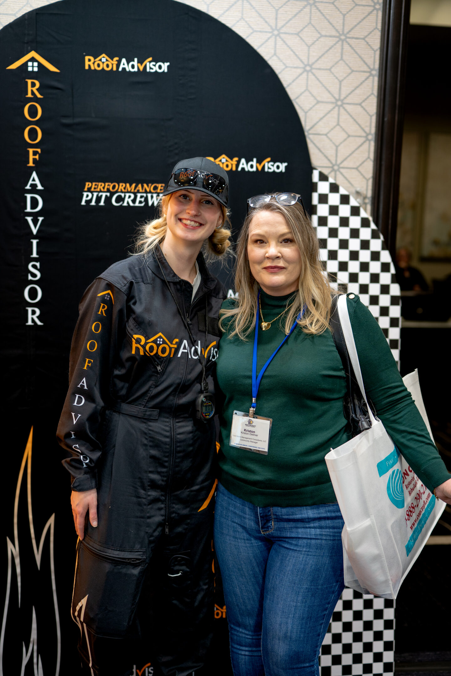 Two women pose at a Roof Advisor event, CAI2026018. One wears a branded jumpsuit. The other wears jeans and a green sweater. Both are smiling.