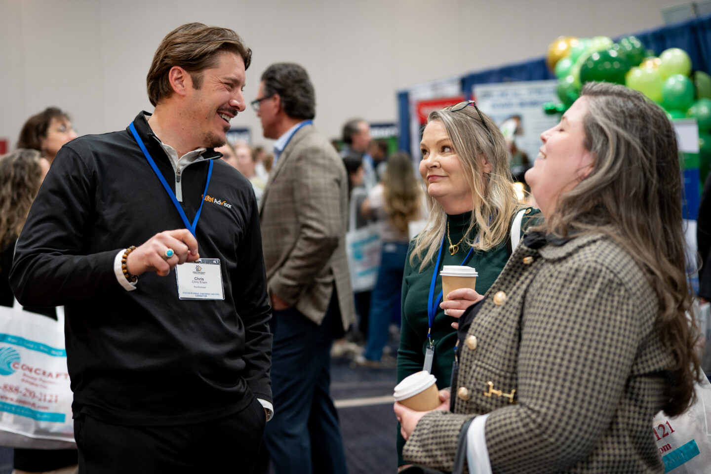 CAI2026019: Three people are having a conversation at a conference. One man and two women are dressed professionally and holding coffee, smiling.