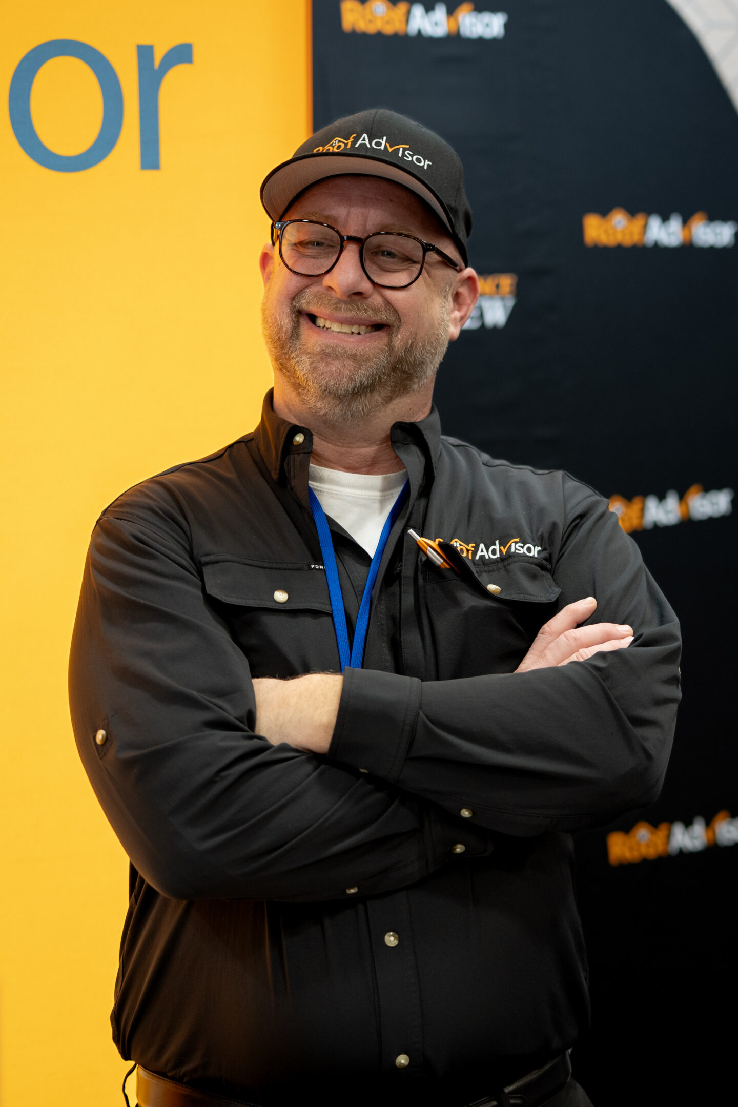 CAI2026020: Man wearing a Roof Advisor cap and black shirt smiles with crossed arms. He's at a conference, showcasing Roof Advisor services.