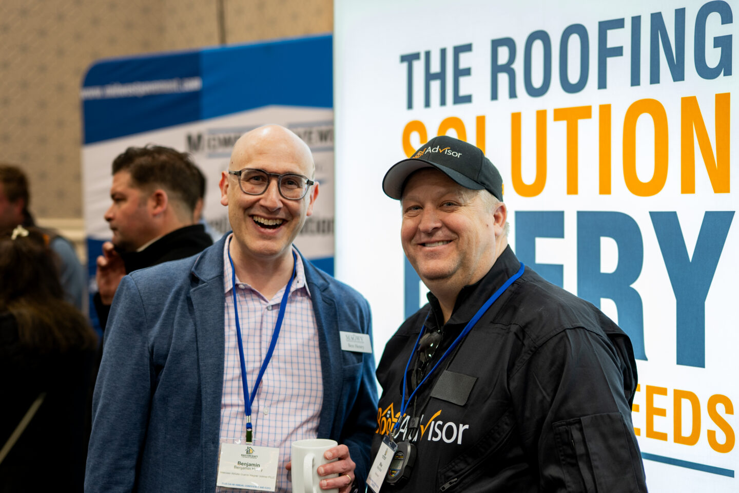 Two smiling men at a roofing advisor booth, CAI2026021.jpg. They are at a conference. One man is wearing glasses and a blue jacket. The other is in a black cap.