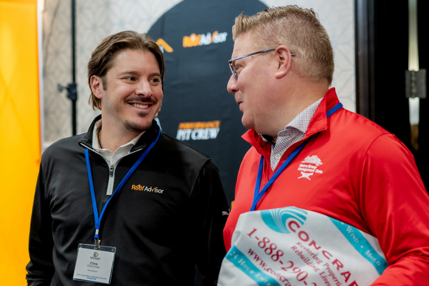 Two men at a trade show, CAI2026022, one wearing a Roof Advisor shirt and the other a Metro Green shirt, discussing concrete restoration and rebuilding properties.