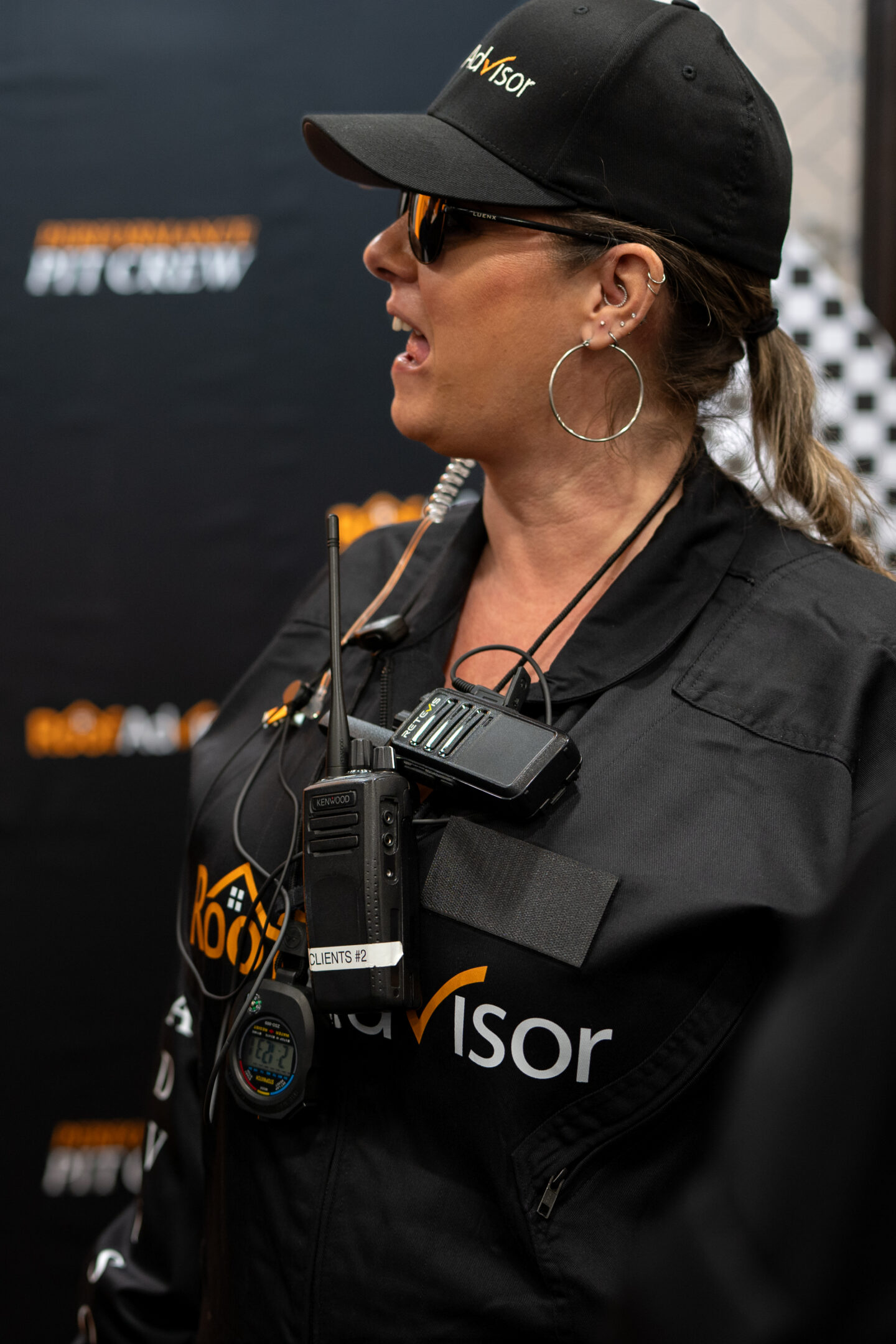 CAI2026026: Woman in uniform with 'Advisor' cap, sunglasses, and radio equipment. Pit crew member at event, showcasing team communication and branding.
