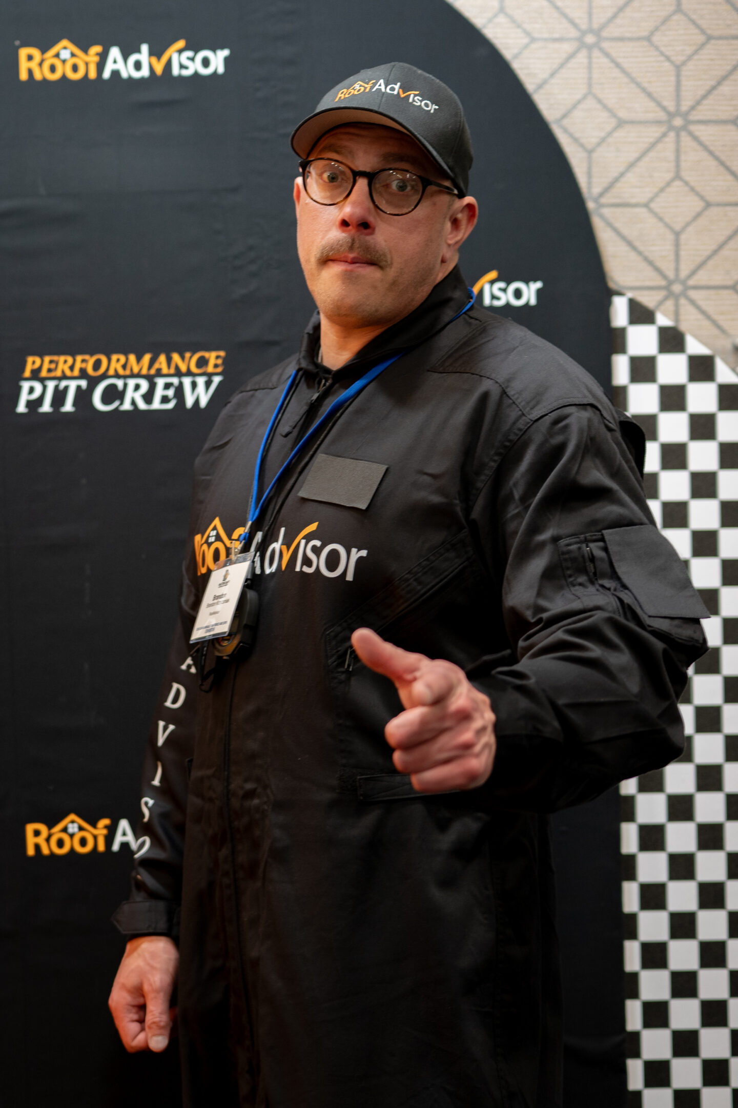 Roof Advisor representative in a black jumpsuit and cap, pointing at the camera. CAI2026028 highlights the advisor in the performance pit crew.