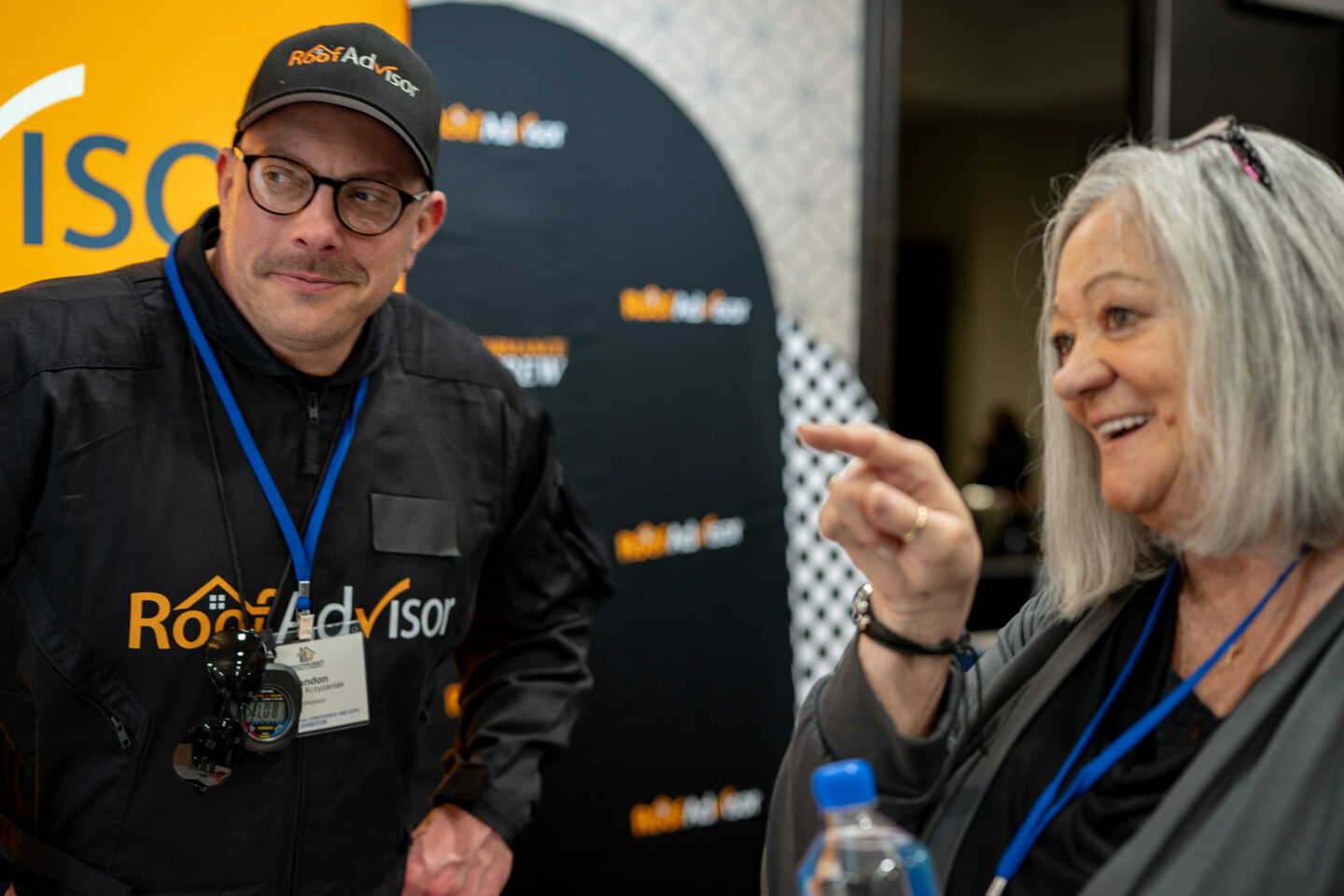 CAI2026029: Man in Roof Advisor hat and woman smiling and pointing. Roofing industry event. Learn more about roofing solutions and expert advice.