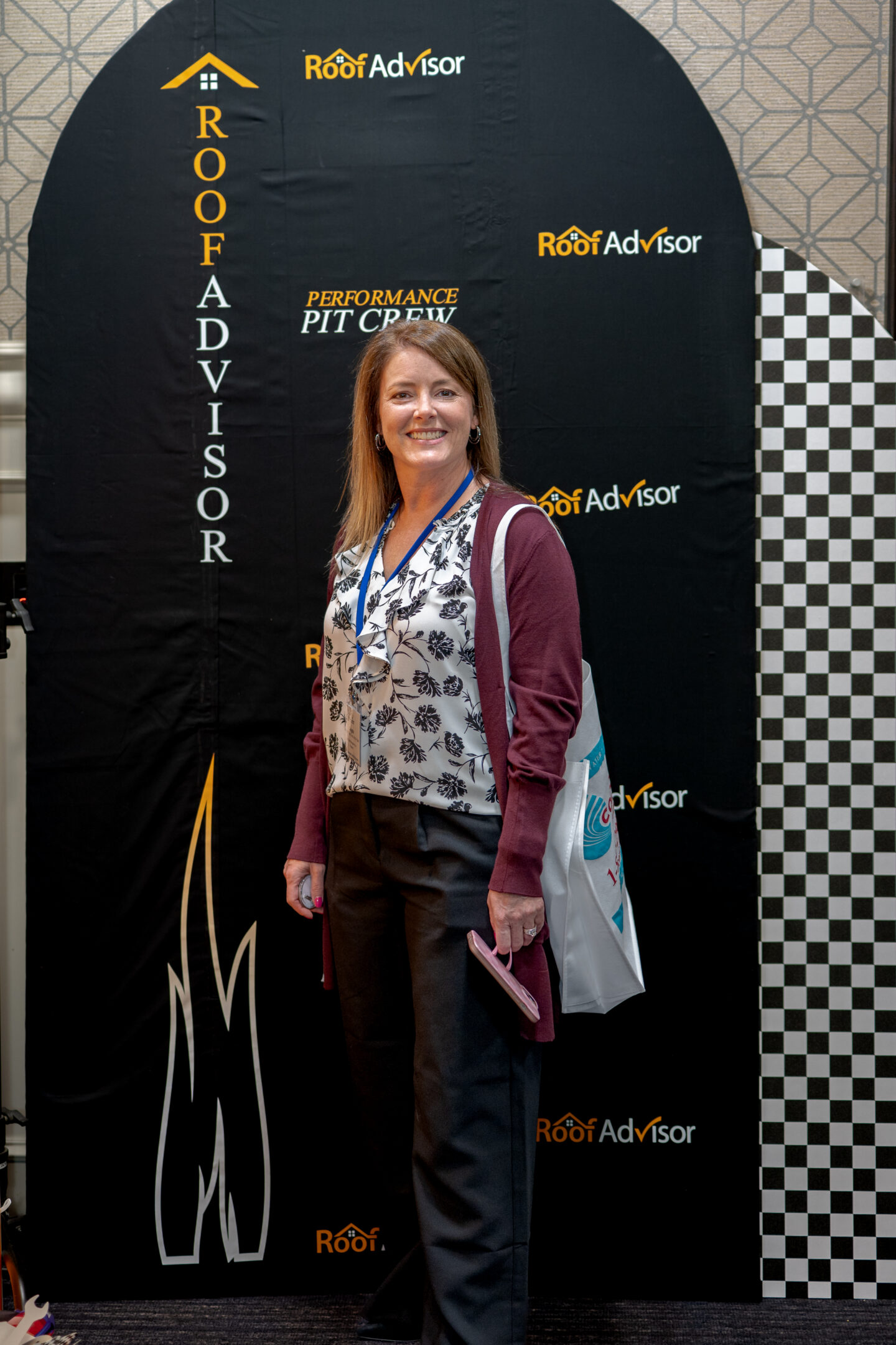 CAI202603: A smiling woman stands in front of a Roof Advisor backdrop. The banner displays roofing company branding and logos, promoting their advisor services.