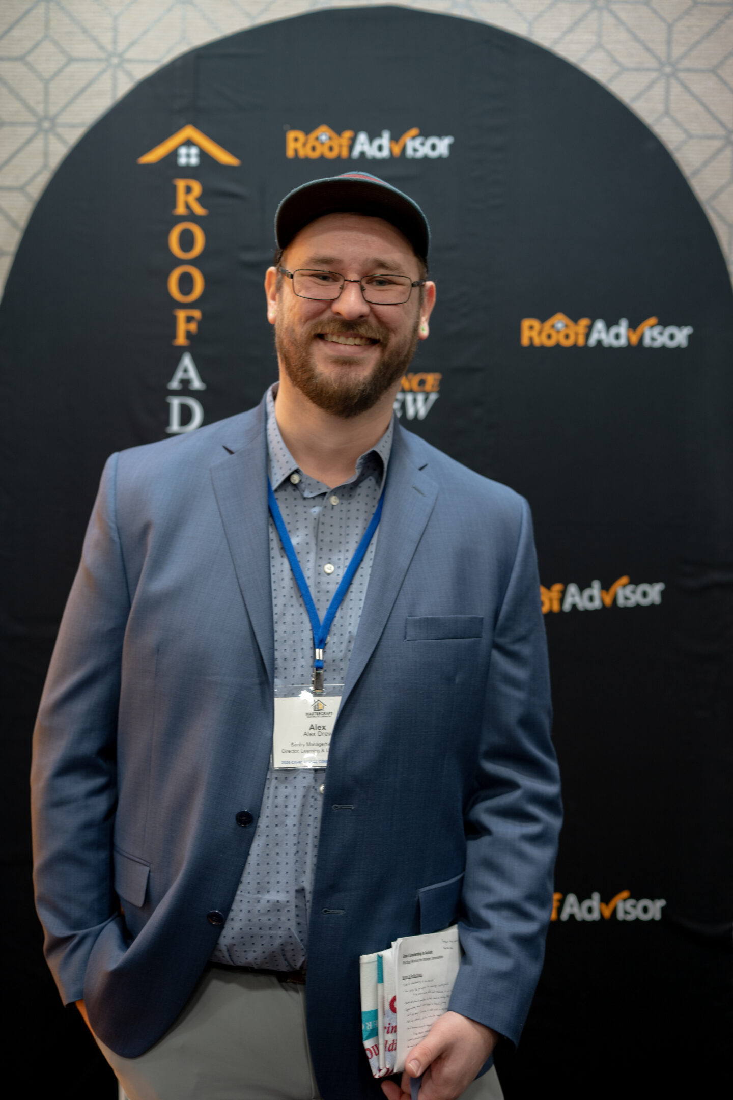 CAI2026030: Man in suit with Roof Advisor backdrop. He is at a roofing industry event, holding papers. He looks friendly and approachable.