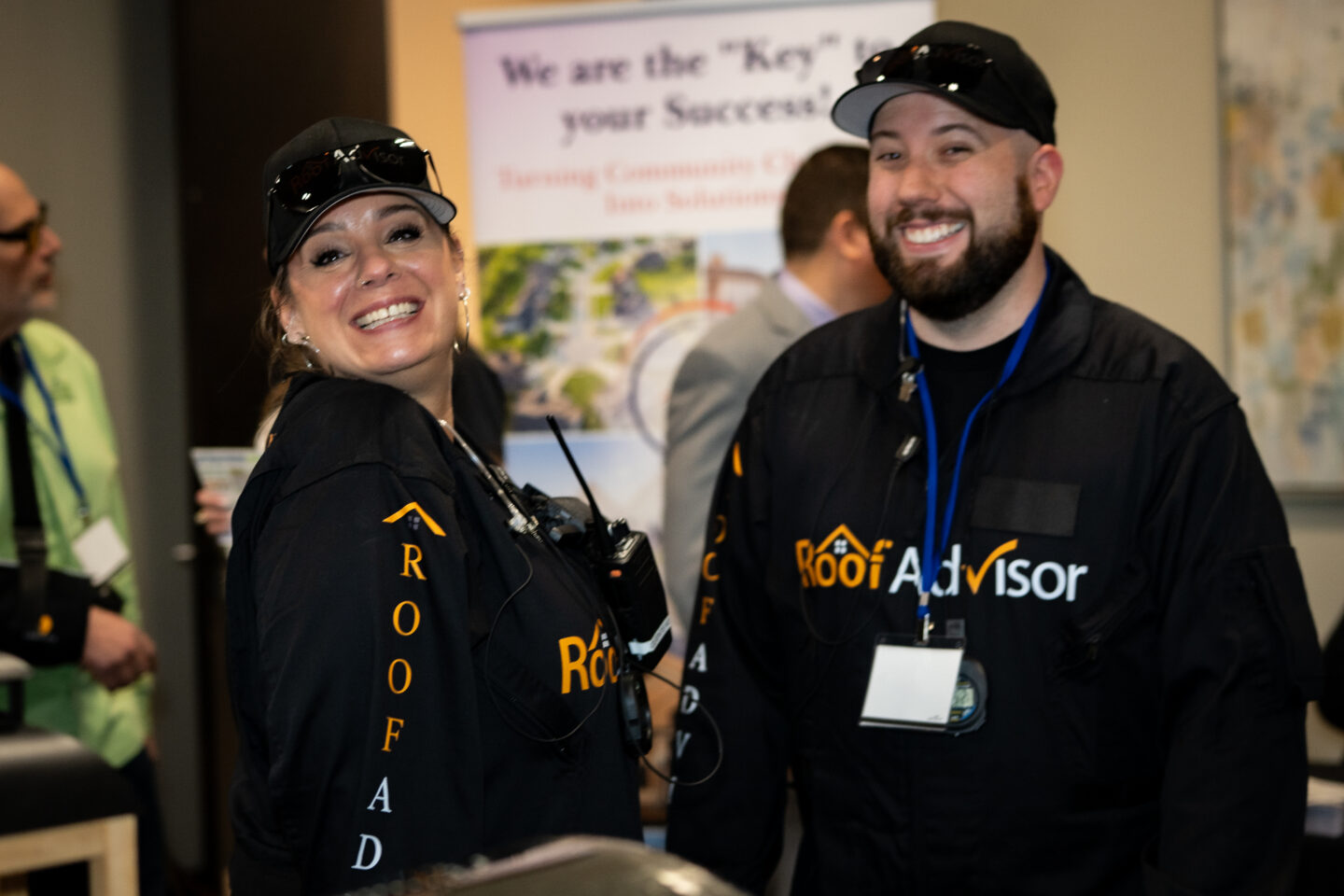 Two smiling representatives from Roof Advisor, CAI2026032, wearing branded jackets. A woman with a walkie-talkie and a bearded man are promoting roof solutions.