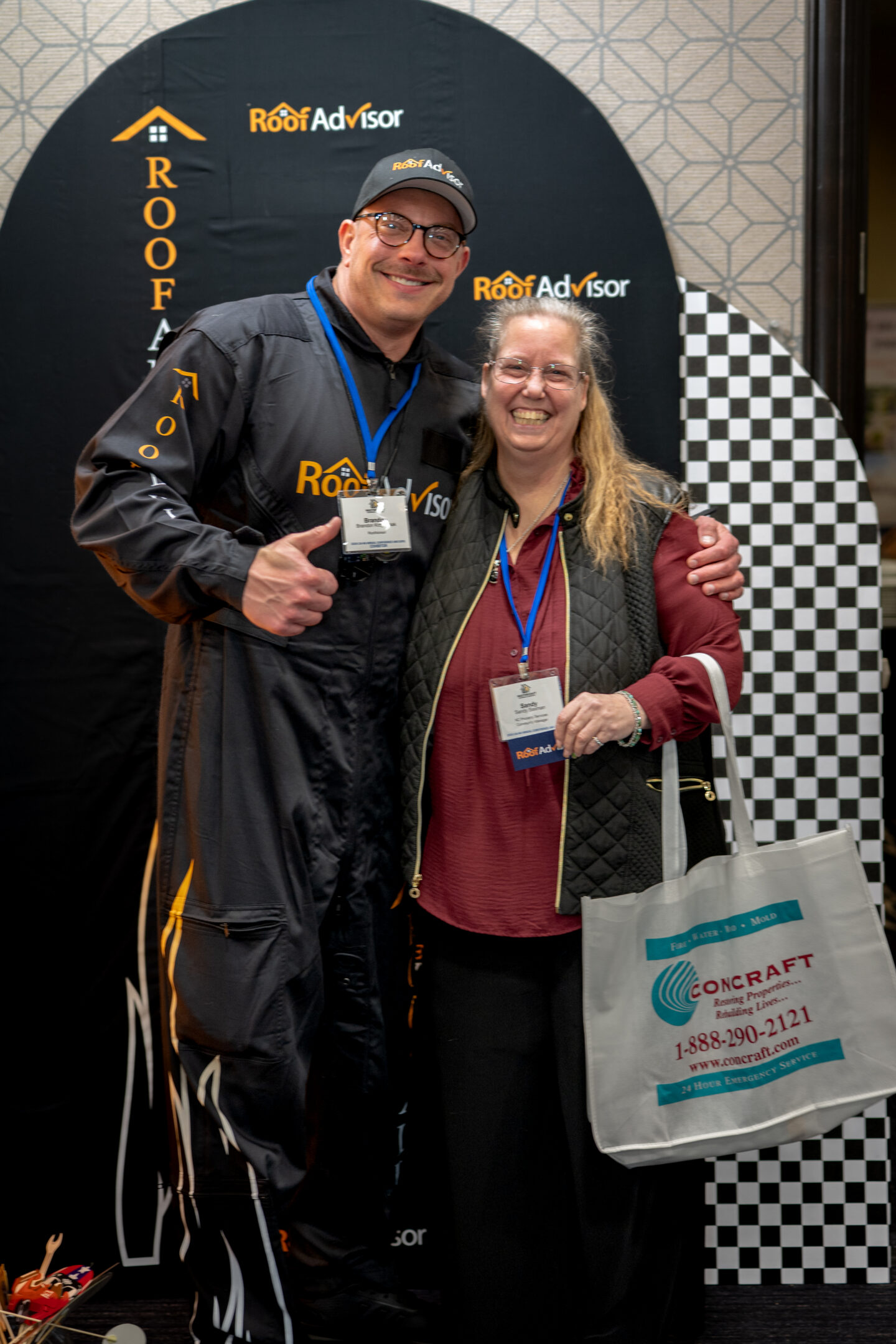 Roof Advisor CAI202604: Two smiling people pose in front of a Roof Advisor backdrop. One wears a branded jumpsuit, the other holds a Concraft bag.