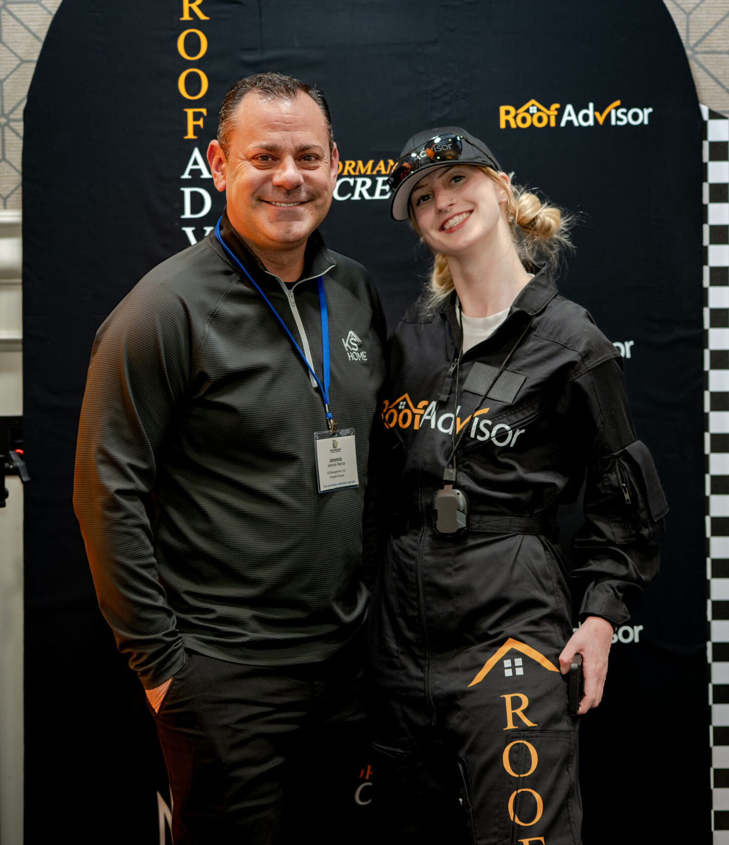 Two individuals, one in a RoofAdvisor-branded jumpsuit, pose at an event. CAI202606 showcases a roofing company's presence and team spirit. Photo op.