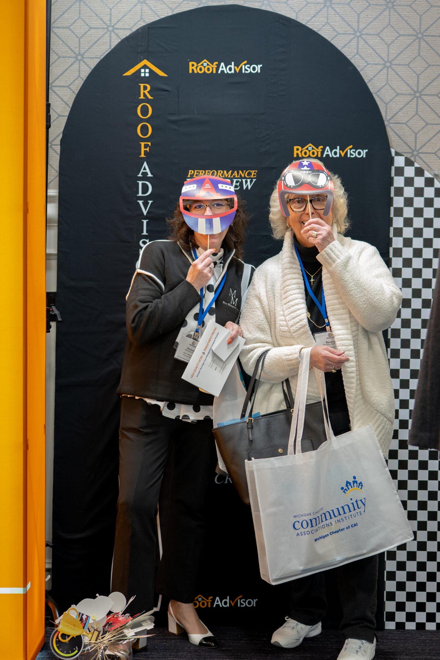 Two women wear racing masks at a Roof Advisor booth, CAI202607. They hold sticks to their faces, with Roof Advisor branding in the background.