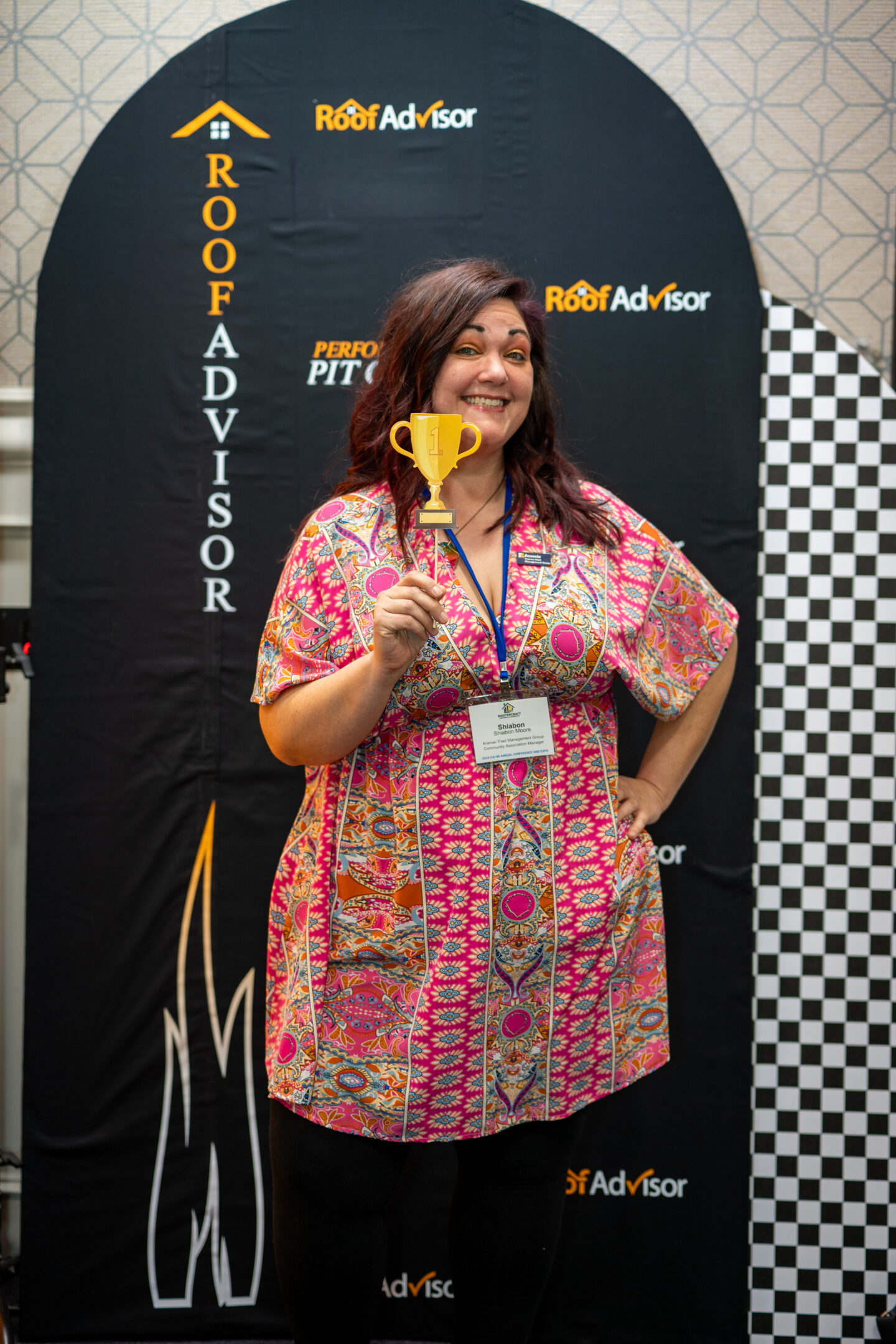 CAI202608: Woman smiles, holding a gold trophy in front of a Roof Advisor banner. She wears a colorful dress and a name tag, celebrating a win.