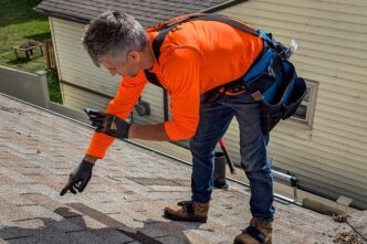 Home: Roofer inspecting roof shingles for damage. Roofing contractor wearing safety harness and tool belt.
