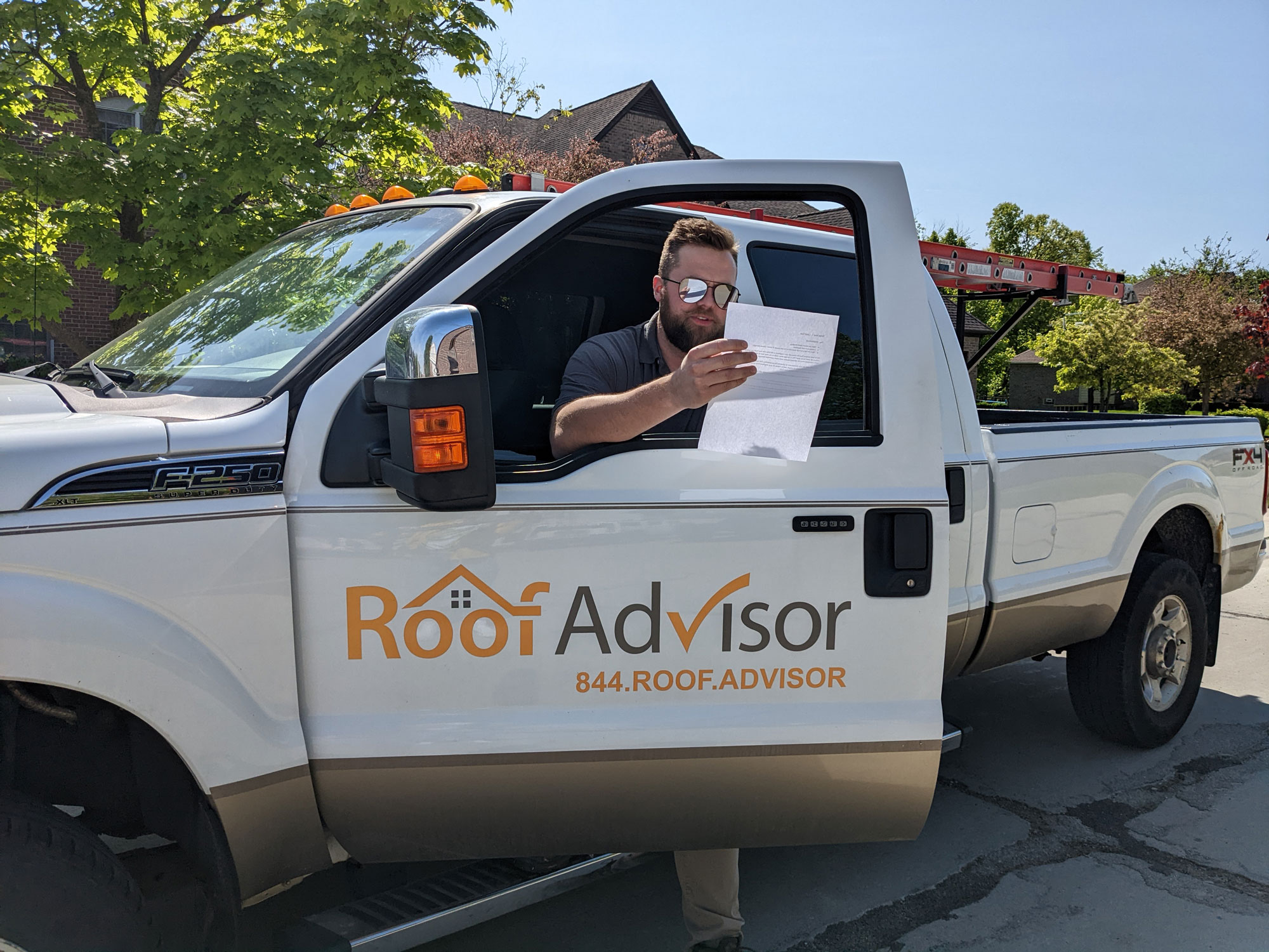 Roof Advisor at job site in truck. Man in truck reads paperwork. Ladder in truck bed. Roof Advisor logo and phone number on truck door.