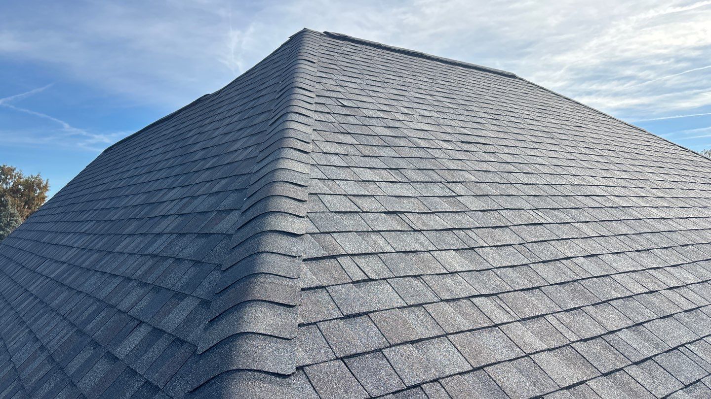 Gray shingle roof with a peaked ridge under a partly cloudy blue sky. After Replacement