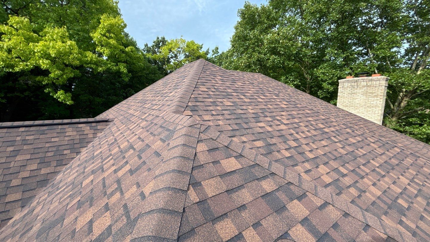 Brown asphalt-shingle roof with multiple ridges, surrounded by trees and a brick chimney