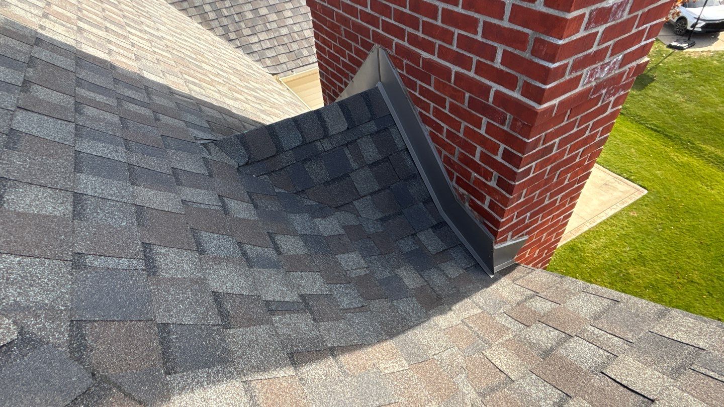 Close-up of a brick chimney with newly installed asphalt shingles and metal flashing on a residential roof.