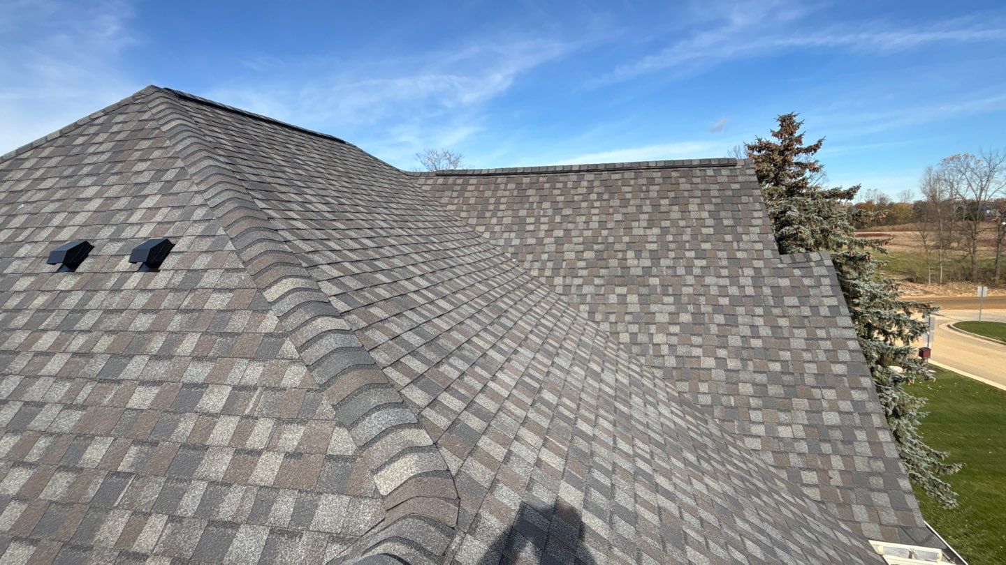 Gray and brown asphalt shingles cover a large, multi-section roof under a clear blue sky