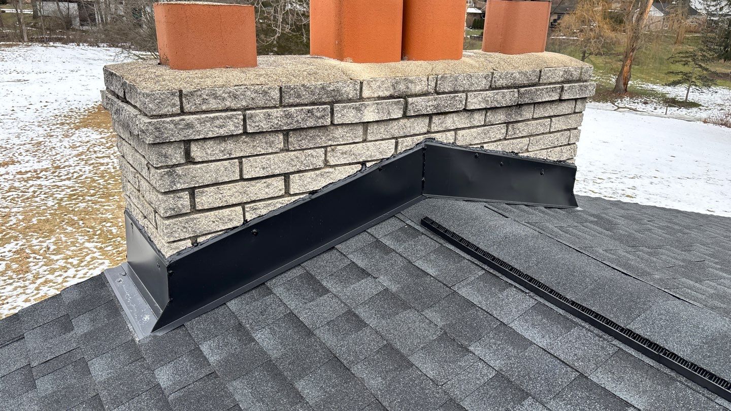 Close-up view of a brick chimney with newly installed black metal flashing at the base on a dark shingled roof.
