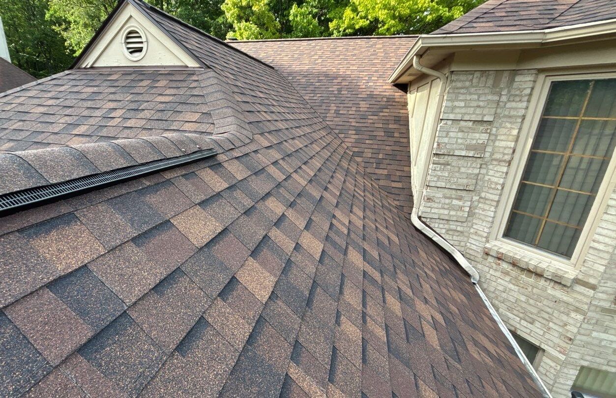 Close-up of overlapping asphalt shingles on a multi-section residential roof beside a brick dormer