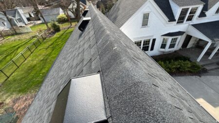 A high angle view of a gray asphalt shingle roof on a residential home with black roof vents visible against the backdrop of a white house and green lawn