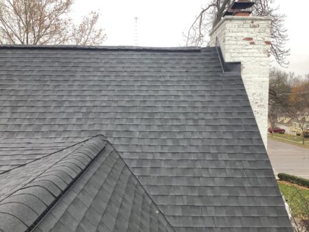 Close up on a dark charcoal shingled roof featuring a prominent white painted brick chimney set against a suburban street view with autumn bare trees