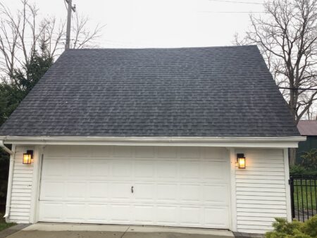 Exterior view of a detached garage featuring a new dark asphalt shingle gable roof with white horizontal siding and two glowing carriage-style wall lamps