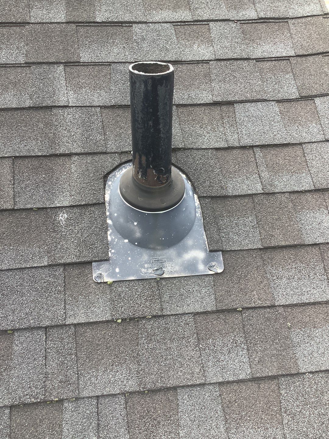 Weathered asphalt shingles surrounding a metal roof vent pipe with an IPS Waterlite flashing base that shows signs of wear and mineral deposit buildup.