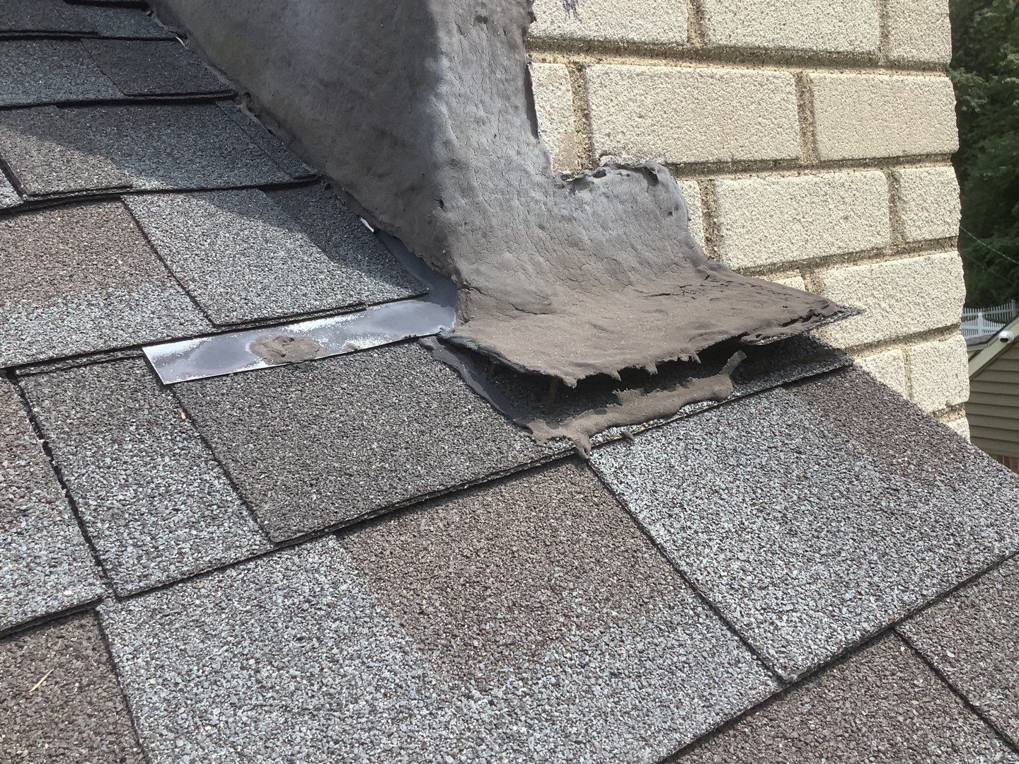 Close-up of a damaged roof area in Dearborn showing worn asphalt shingles and thick gray sealant applied poorly along the intersection with a brick chimney.