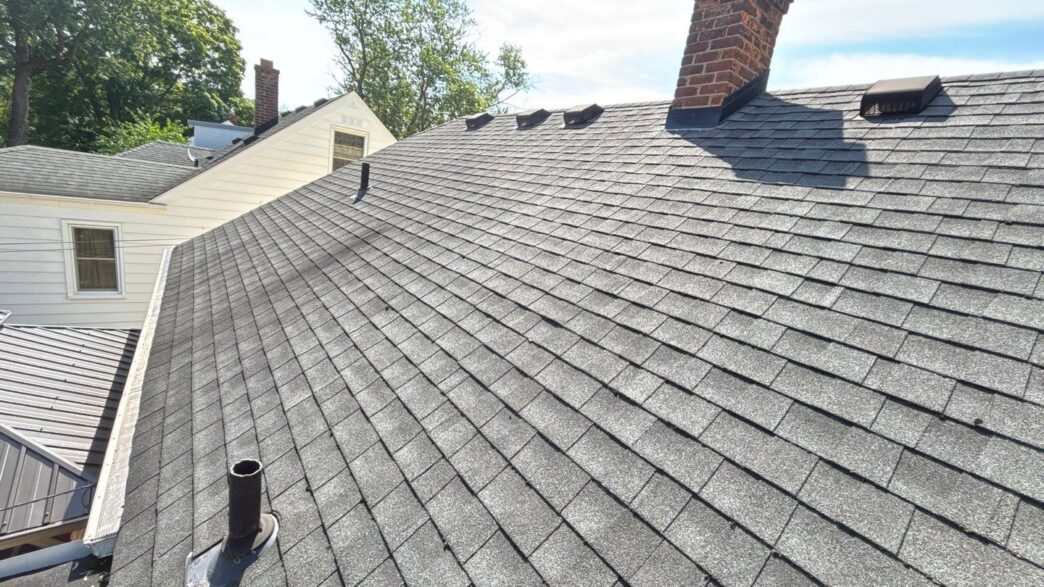 Roof with gray asphalt shingles, chimney, and vent pipes on a sunny day