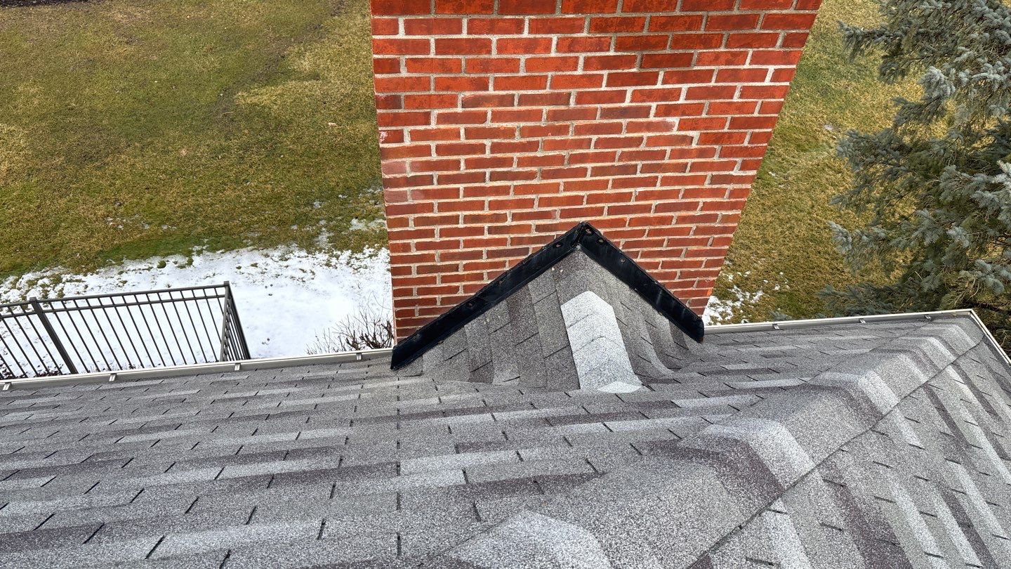 Worn shingled roof with dark streaks and discoloration around the base of a red brick chimney, overlooking a fenced yard.