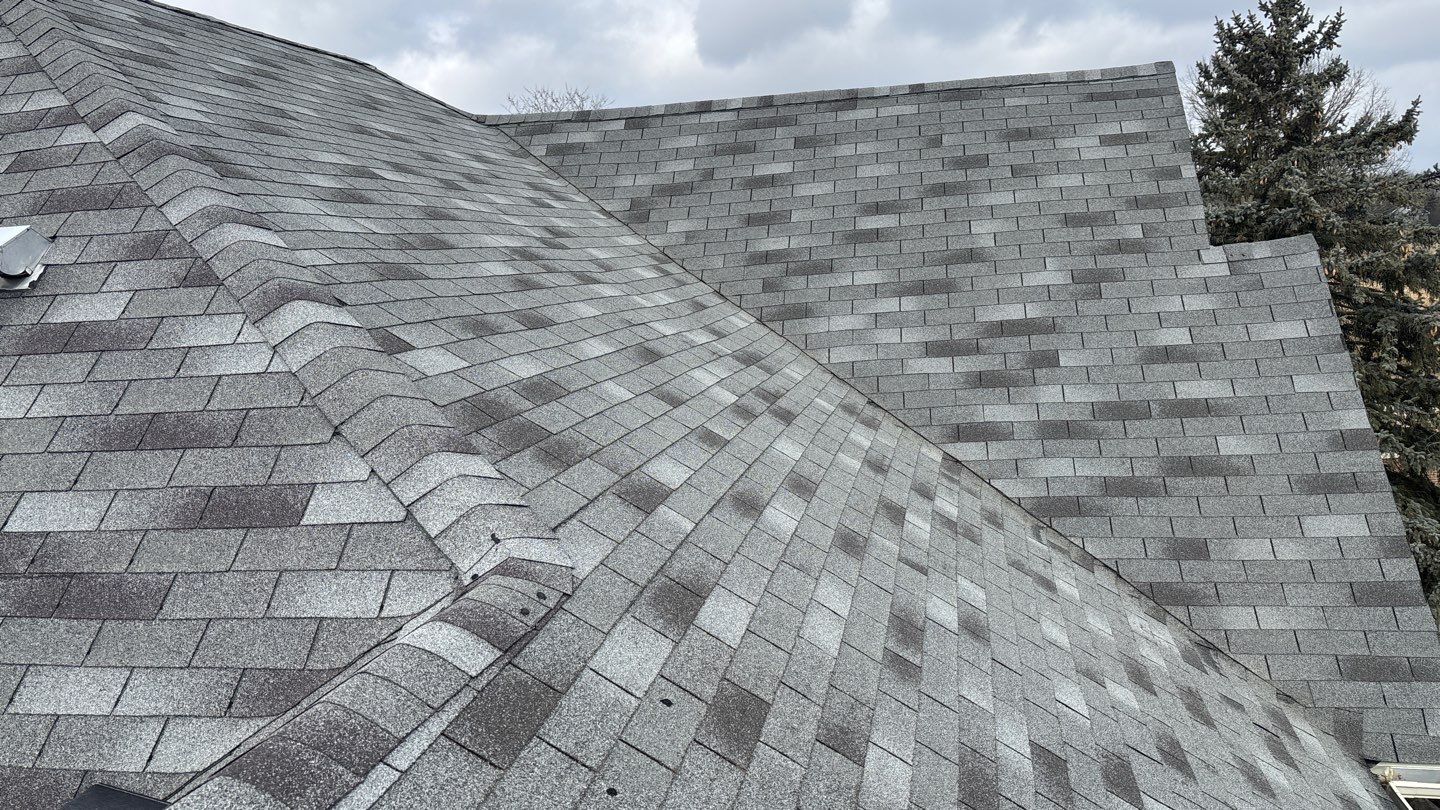 Gray asphalt shingle roof with prominent valleys and ridges, set against a cloudy sky and a tall evergreen tree nearby.