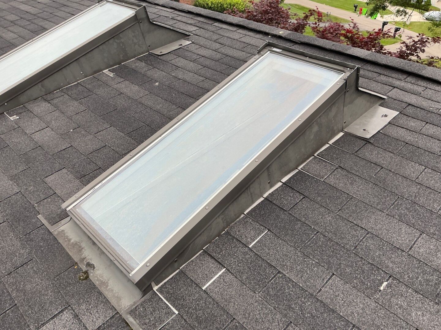 Rectangular skylight with metal flashing installed on a dark shingled roof, garden plants visible in background.