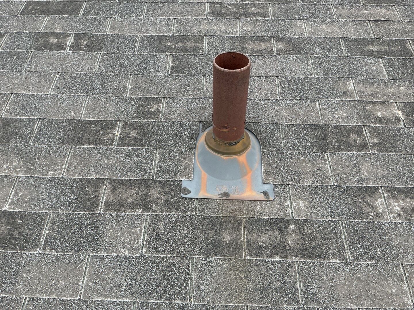 A close-up view of a roof vent pipe with worn flashing surrounded by gray asphalt shingles showing signs of weathering.