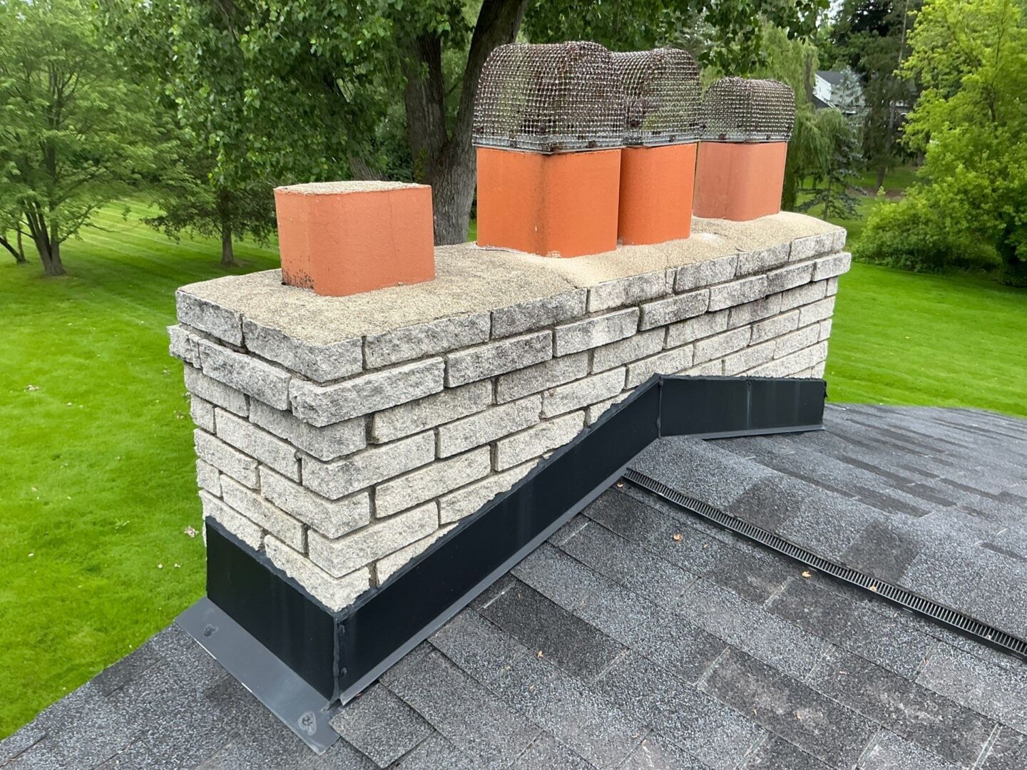 A brick chimney with three orange chimney flue liners sits on a shingled roof, surrounded by green grass and trees.