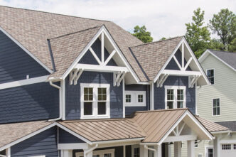 Belmont Colonial Slate Shingles on a home. Close-up of a blue house with gray shingles and white trim, showcasing roofing detail. Sun cover uv resistant granules that help reduce energy costs.