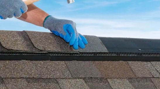 Integrity roofing: Installing hip and ridge accessories and shingles. A gloved hand nails shingles to a roof under a blue sky, showcasing roofing detail and craftsmanship.
