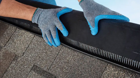 Integrity roof system: Installing the ridge ventilation on shingles. Hands in blue-tipped gloves secure black ventilation strip to a roof. Roofing with proper attic airflow.