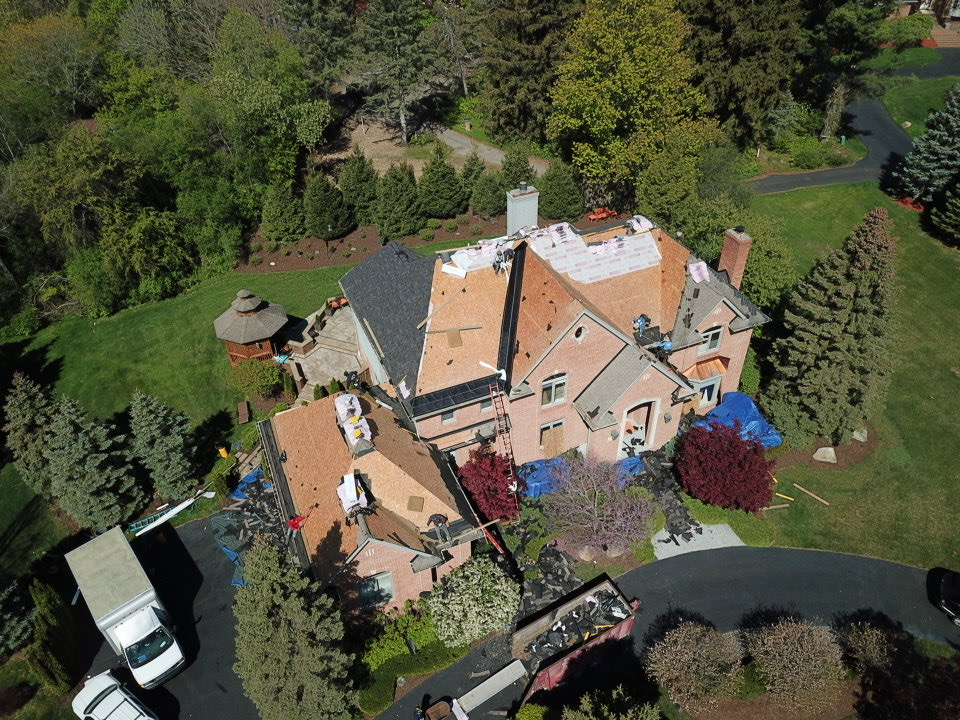 Testimonial image showcasing a complete roofing system replacement project. Aerial view of a house with a partially replaced roof, trucks, and workers. Roofers at work doing an outstanding job.