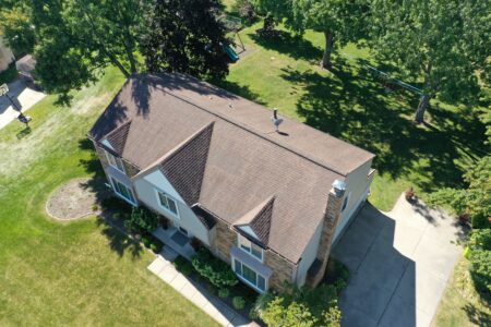 Before roof replacement in Novi, MI. View of house with brown roof, green lawn, and trees. Get to Know RoofAdvisor for roof replacement solutions.