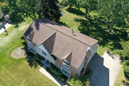 Before roof replacement in Novi, MI. View of house with brown roof, green lawn, and trees. Get to Know RoofAdvisor for roof replacement solutions.