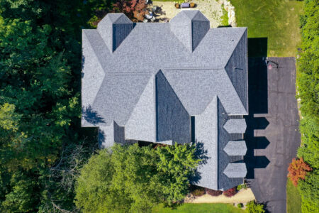 Aerial view of a finished roof replacement on a large house surrounded by trees and lawn. Gray asphalt shingles cover the complex roof structure.