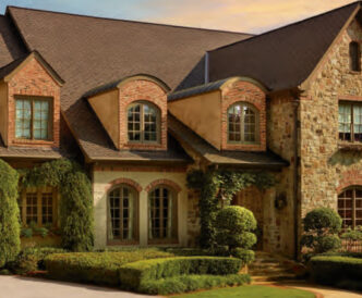 Designer GAF Grand Sequoia example of laminate shingles in Mesa Brown. Mimics wood shakes and cedar shakes. Upscale home's curb appeal with stone and brick exterior, arched windows, and manicured landscaping.