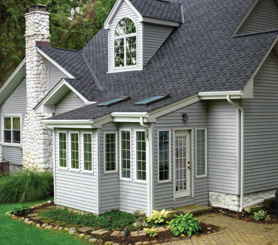 GAF Timberline Grand Sequoia Designer roof example. Gray asphalt shingle roof on a gray house with white trim, stone chimney, and a sunroom.