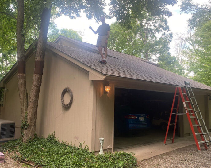 After Canton roof replacement: Man standing on garage roof giving a thumbs up, ladder, garage door open with car inside. Landscaping and trees visible.